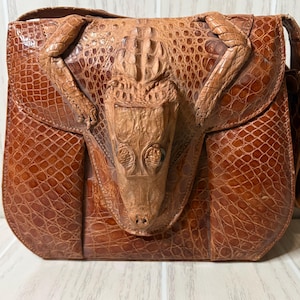 May include: A brown leather handbag with a crocodile head design on the flap. The bag has a shoulder strap and a textured, scaly pattern. The crocodile head is intricately detailed, adding a unique touch to the accessory.