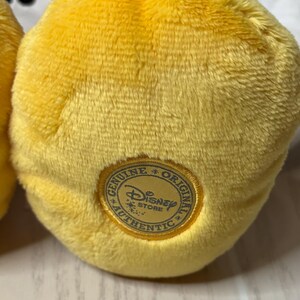 May include: Close-up of a plush yellow toy with a textured surface. A gold-colored circular patch with the words "Genuine Original Authentic Disney Store" surrounds the Disney logo.