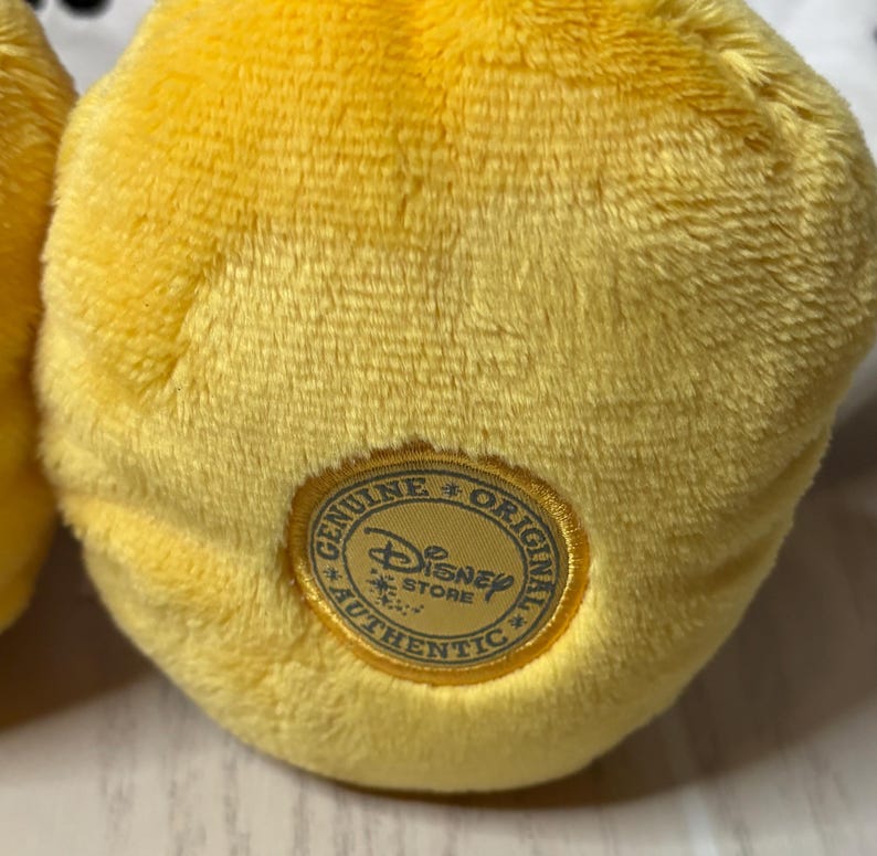 May include: Close-up of a plush yellow toy with a textured surface. A gold-colored circular patch with the words "Genuine Original Authentic Disney Store" surrounds the Disney logo.