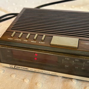 Puede incluir: Un radio reloj digital FM/AM Emerson vintage. El radio reloj tiene un acabado de madera marr&oacute;n con botones para hora, minuto, tiempo, despertar y dormir. La pantalla muestra la hora en rojo. La radio tiene un bot&oacute;n de repetici&oacute;n y un cable de alimentaci&oacute;n negro.