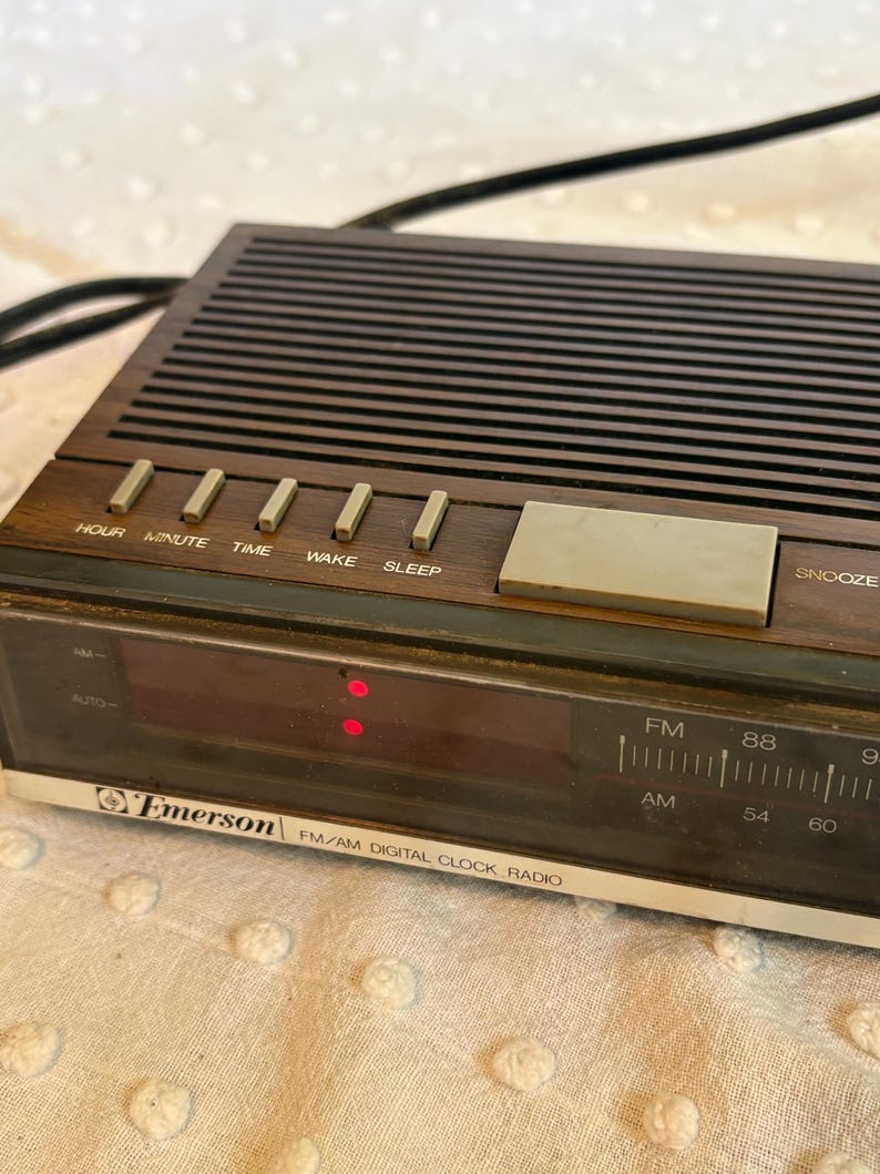 Puede incluir: Un radio reloj digital FM/AM Emerson vintage. El radio reloj tiene un acabado de madera marr&oacute;n con botones para hora, minuto, tiempo, despertar y dormir. La pantalla muestra la hora en rojo. La radio tiene un bot&oacute;n de repetici&oacute;n y un cable de alimentaci&oacute;n negro.