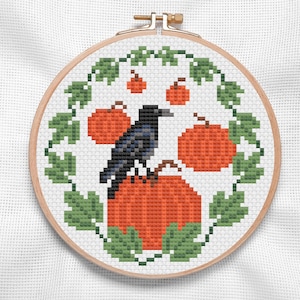 May include: Cross stitch pattern featuring a black crow perched on a pumpkin with a wreath of green leaves and orange pumpkins around it.