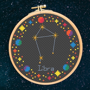 May include: A black and white embroidery hoop with a colorful cross-stitch design of the Libra constellation. The design features a stylized scale with stars and the word "Libra" in white text.