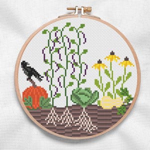 May include: Cross-stitch embroidery in a wooden hoop. The design features a garden scene with a pumpkin, a crow, purple beanstalks, a cabbage, and yellow flowers. The roots of the plants are visible in the soil.