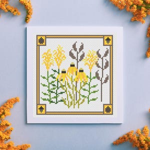Puede incluir: Un diseño de punto de cruz de flores amarillas y hojas marrones sobre un fondo blanco, enmarcado con un borde amarillo y marrón. La obra de arte está en una tarjeta cuadrada blanca, con flores naranjas en las esquinas.