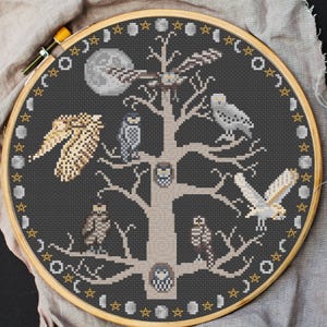May include: Cross-stitch embroidery of owls perched on a tree, set against a dark gray background. The design includes a full moon, stars, and phases of the moon, all within a wooden embroidery hoop. The owls are in various shades of brown, gray, and white.