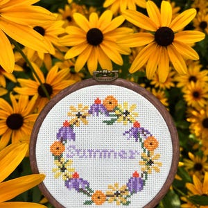 May include: A cross-stitch embroidery hoop with a wooden frame. The design features a floral wreath in yellow, purple, and orange, with the word "Summer" stitched in the centre. The background is filled with yellow flowers.