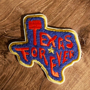 Op de afbeelding: Een blauwe viltpatch in de vorm van de staat Texas met de woorden "Texas Forever" geborduurd in rood. Een gouden rand omringt de patch.
