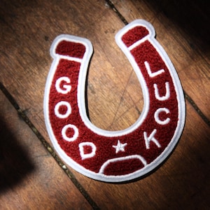 May include: A horseshoe-shaped patch with a textured deep red surface and a white border. The text GOOD LUCK is embroidered in white capital letters along the curve of the horseshoe, with a small white star centered at the bottom of the design.