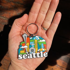 May include: A colorful rubber keychain featuring a stylized skyline of Seattle, Washington. The keychain includes the Space Needle, a Ferris wheel, and the text "seattle".