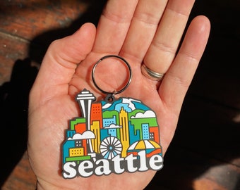 Puede incluir: Un llavero de goma colorido con un horizonte estilizado de Seattle, Washington. El llavero incluye la Space Needle, una noria y el texto "seattle".