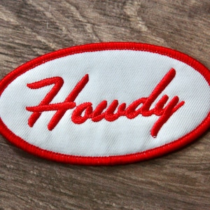 May include: Embroidered patch with the word "Howdy" in red script on a white oval. The oval is outlined with a red border. The patch is likely for clothing or accessories.