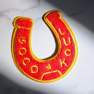 May include: Red felt horseshoe with a yellow outline and the words "GOOD LUCK" embroidered on it.