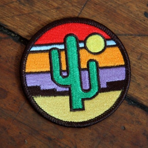 May include: A round embroidered patch with a green cactus in the center, a sunset with orange, yellow, and red stripes, and a yellow sun in the upper right corner.