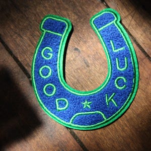 May include: A horseshoe-shaped patch made of blue felt with a bright green embroidered border. The text GOOD LUCK is stitched in green thread along the curve of the patch, with a small green star positioned between the letters D and K.