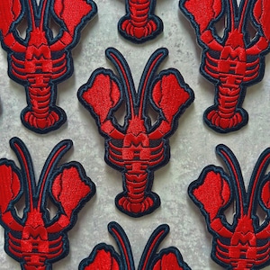 Könnte beinhalten: Rot gestickte Hummer-Patches mit marineblauer Umrandung. Die Patches weisen detaillierte Nähte auf, die die Scheren, Körpersegmente und Antennen des Hummers hervorheben. Diese Patches eignen sich für Kleidung, Taschen oder andere Stoffartikel.
