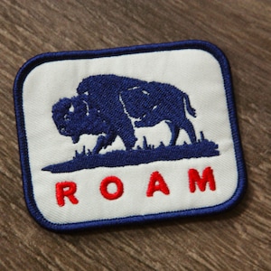May include: Embroidered patch featuring a blue bison silhouette on a white background, with the word "ROAM" in red block letters. The patch has a blue border and is likely for clothing or accessories.