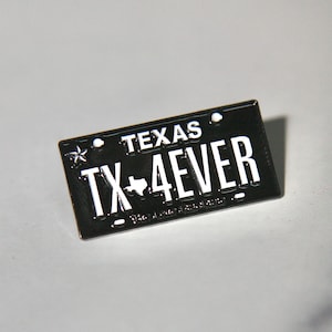 Pode incluir: Um alfinete preto em forma de placa de carro com o texto "TEXAS" no topo e "TX 4EVER" em letras brancas. Uma estrela branca e o contorno do Texas também são visíveis no alfinete.