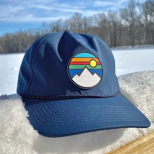 May include: A navy blue baseball cap with a colorful mountain and sun graphic patch. The cap has a curved brim and a white stitching detail around the edge.
