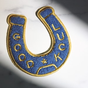 May include: A blue horseshoe-shaped patch with gold trim and the words "GOOD LUCK" embroidered in gold. The patch has a textured appearance and is likely made of fabric, possibly for application on clothing or accessories.