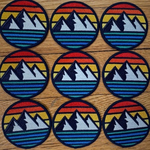 May include: Nine embroidered patches with a mountain range design. Each patch features a sunset with orange, yellow, and red hues, a blue lake, and a white mountain range. The patches are circular and have a black border.