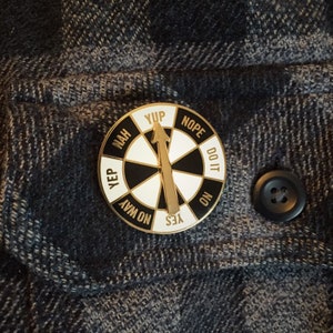 May include: A gold-colored pin with a black and white wheel that has the words "Yup", "Nope", "Do It", "No", "Yes", "No Way", "Yep", and "Nah" printed around the edge. The wheel has a gold arrow pointing to the word "Yes".