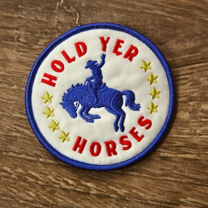 May include: Embroidered patch with a blue cowboy riding a blue horse, surrounded by the words "HOLD YER HORSES" in red, and yellow stars. The patch has a white background with a blue border.