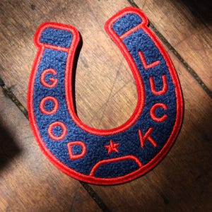 May include: A horseshoe-shaped embroidered patch with a textured dark blue surface and a bright red border. The words GOOD LUCK are stitched in red thread along the curve of the patch, with a small red star separating the words at the bottom.