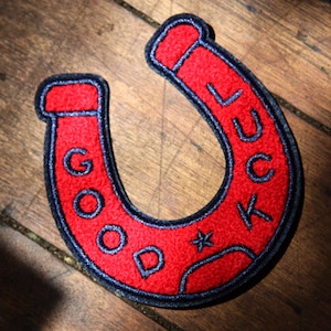 May include: A red horseshoe-shaped patch with a dark blue embroidered border and the text GOOD LUCK stitched in dark blue thread. A small dark blue star is positioned between the words GOOD and LUCK on the red felt-like surface of the patch.