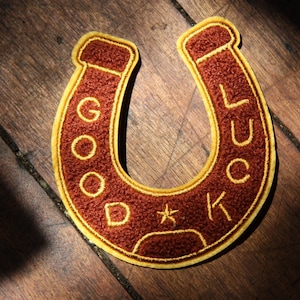 May include: A horseshoe-shaped embroidered patch with a textured dark rust-colored surface and a bright yellow border. The text GOOD LUCK is stitched in yellow thread along the curve, with a small five-pointed star centered at the bottom.
