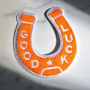 May include: Orange horseshoe-shaped patch with white embroidered text reading "GOOD LUCK" and a star. The patch has a white border and a textured, fuzzy appearance. Ideal for adding to clothing or accessories.