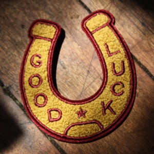 May include: A horseshoe-shaped embroidered patch featuring a textured gold-colored surface with a dark red border. The text GOOD LUCK is stitched in dark red thread along the curve of the patch, with a small red star centered at the bottom.