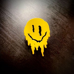 May include: A yellow smiley face pin with black outline, dripping with yellow paint. The pin is on a dark brown surface.