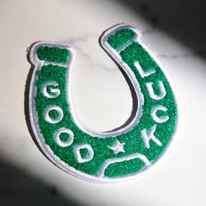 May include: Green felt horseshoe patch with white stitching and the words "GOOD LUCK" embroidered around the horseshoe.