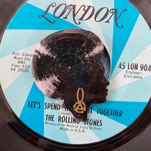 Può includere: Un disco in vinile bianco e nero con una silhouette di una testa di donna con un orecchino dorato. Il disco è intitolato "Let's Spend It Together" dei Rolling Stones. L'etichetta è blu e bianca con il testo "45 LON 904" e "Engineer: Glyn Johns".