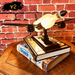 May include: A unique airplane-shaped lamp made from a repurposed bottle, with black wings and engines. The lamp sits atop two books, one titled "World at Arms" and the other "Chronicle". The background is a red brick wall. The text "v2" is in the upper left corner.