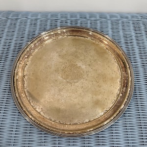 May include: A round silver-toned metal tray with a polished rim and a tarnished, weathered center surface. The center of the tray features an intricate, engraved monogram consisting of overlapping cursive letters, creating a decorative focal point.
