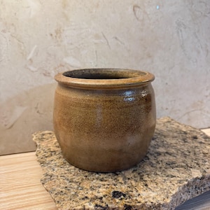 May include: A small, round, handcrafted ceramic pot with a wide opening. The pot has a speckled, earthy brown glaze with darker brown accents around the rim. The pot is sitting on a rough, textured stone surface.