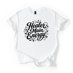 Könnte beinhalten: Ein weißes Kurzarm-T-Shirt mit schwarzem Schriftzug Healer Main Energy in einer stilisierten Schreibschrift. Der Text ist von dekorativen schwarzen, geschwungenen Ranken- und Blattillustrationen in der Mitte des Kleidungsstücks umgeben.