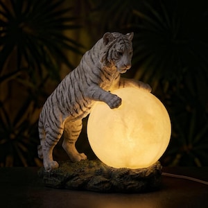 May include: A white tiger figurine with black stripes standing on a textured green base. The tiger has one paw resting on a large, spherical, glowing lamp that emits a warm, soft yellow light, illuminating the tiger's body and the surrounding area.