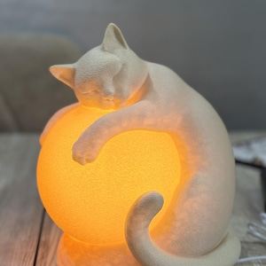 May include: A cream-coloured cat figurine lamp with a textured surface, featuring a cat curled around a glowing, spherical orange light source. The lamp has a solid base and emits a warm, bright light from the centre sphere, illuminating the cat.