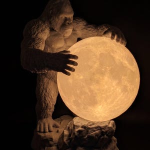 Puede incluir: Una figura de gorila blanco sobre una base rocosa, sosteniendo una gran lámpara de luna esférica y brillante. El gorila tiene una textura de pelaje detallada y rasgos musculosos, mientras que la esfera lunar iluminada muestra una superficie con cráteres.