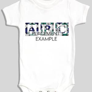 May include: White baby onesie with short sleeves. The front features the letters "ABC" in a square design with a floral pattern in blue, green, and white. Below the letters, the text reads "PLACEMENT EXAMPLE".