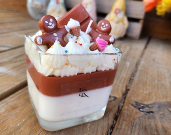 May include: A square glass candle with layers of white and brown wax, topped with white frosting, chocolate, and gingerbread men. The candle is decorated with sprinkles and miniature ice cream cones. The candle sits on a wooden surface.