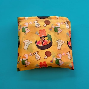 May include: Folded yellow reusable shopping bag featuring a pattern of cartoon ducks, mushrooms, and a bowl of food. The ducks wear green overalls and hold ladles. The bag is on a blue background.