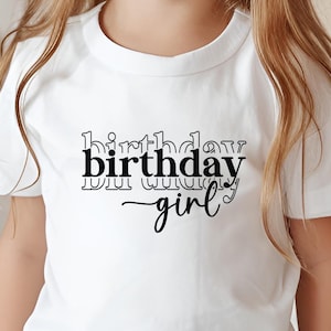May include: A white t-shirt with the words "birthday girl" in black script. The shirt is being worn by a young child. The child is standing in front of a wooden surface.