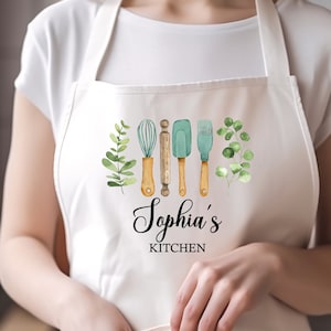 May include: White cotton apron with a pocket, featuring a watercolour design of kitchen utensils and green leaves. The text "Sophia's Kitchen" is printed in black cursive font.