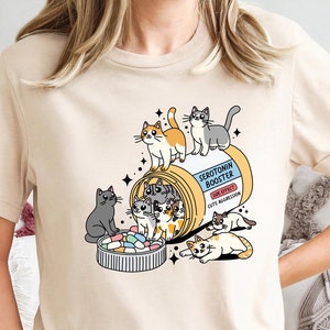 Peut inclure: Un t-shirt beige clair avec un motif représentant une pilule en forme de bouteille étiquetée "Serotonin Booster" avec des chats de dessin animé qui en sortent. Le texte sur la pilule indique "Effet secondaire : Agression mignonne."