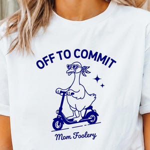 May include: A white short-sleeved t-shirt featuring a dark blue graphic of a goose wearing sunglasses and a bandana while riding a scooter. The text above the goose reads OFF TO COMMIT and the text below the goose reads Mom Foolery.
