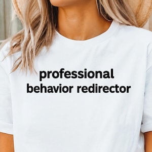 Peut inclure: Un t-shirt blanc à manches courtes avec le texte noir "professional behavior redirector" imprimé dans une police sans empattement simple au centre de la poitrine. Le vêtement présente un col rond classique et une coupe standard.
